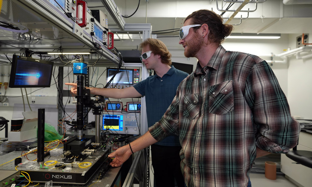 Lindell Williams (left) and Grant Brodnik align an optical fiber with the edge of an integrated photonics chip. Optical fibers act as pipes for light, enabling the light generated on these chips to be collected and routed off the chip for use in experiments and applications. Credit: R. Jacobson/NIST.