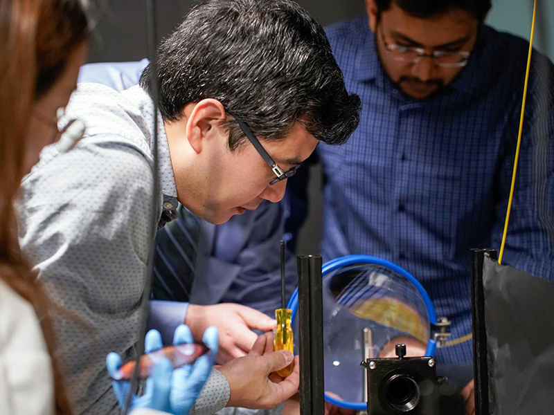 Xingjie Ni (center), Penn State associate professor of electrical engineering, manipulates a conventional telescope lens. In the foreground, Lidan Zhang, electrical engineering graduate student, holds the metalens prototype, which is much smaller, flatter, and more compact. Credit: Jeff Xu/Penn State.