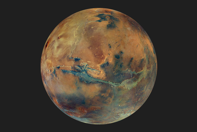 The globe of Mars set against a dark background. The disc of the planet features yellow, orange, blue and green patches, all with an overall muted grey hue, representing the varying composition of the surface. Image credit: ESA/DLR/FU Berlin/G. Michael.