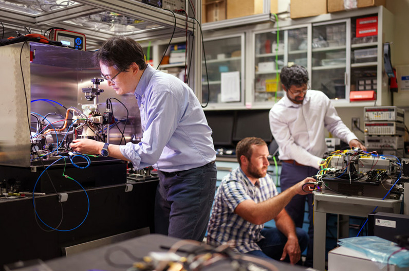 Sandia scientist Jongmin Lee, left, prepares a rubidium cold-atom cell for an atom interferometry experiment while scientists Ashok Kodigala, right, and Michael Gehl initialize the controls for a packaged single-sideband modulator chip. Shoto by Craig Fritz / SNL.