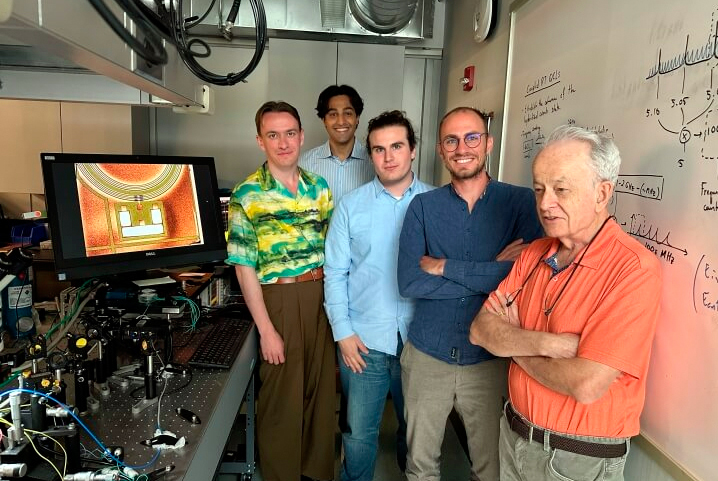 Members of the research team in the Capasso lab. An optical table contains the experimental setup for characterization of the integrated laser sources reported in the paper. The screen shows a microscope image of an active ring resonator based on quantum cascade lasers, used for the demonstration of bright solitons on a laser chip. From left: Dmitry Kazakov, Pawan Ratra, Theodore Letsou, Marco Piccardo, Federico Capasso. 